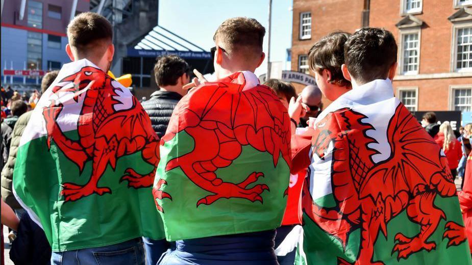 Welsh fans with flags wrapped around them stood in front of the Stadium which can be seen in the background of the image.