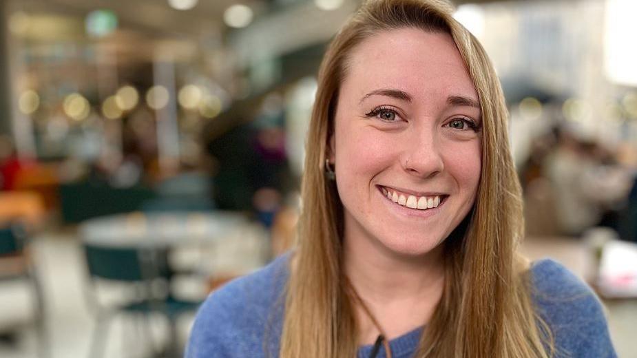 A smiling Rose Steward looks directly at the camera, she is wearing a blue jumper and has long brown hair. 