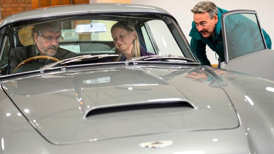 Xhoni dhe Susan janë ulur në makinën ngjyrë gri të argjendtë thupre duke parë panelin e instrumenteve, ndërsa një punonjës i Aston Martin u buzëqesh nga jashtë makinës.