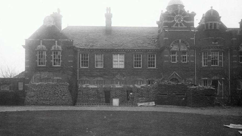 A black and white picture shows a two-storey building with copious amounts of sandbags stacked in front of it.