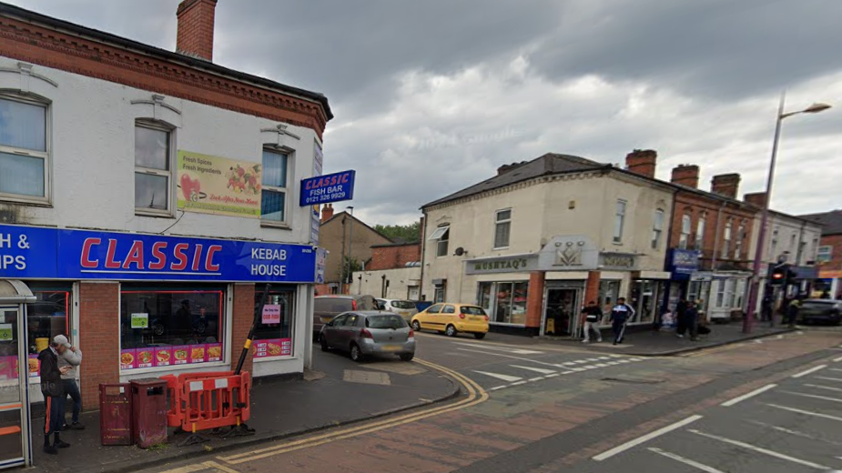 Google street view of a road junction. A fish and chip kebab house can be seen on the left, with a blue sign and a bus shelter outside. On the opposite corner is a south Asian sweet shop with a grey sign. Pedestrians can be seen walking on the pavement and other shops in the distance occupy the ground floors of terraced buildings.