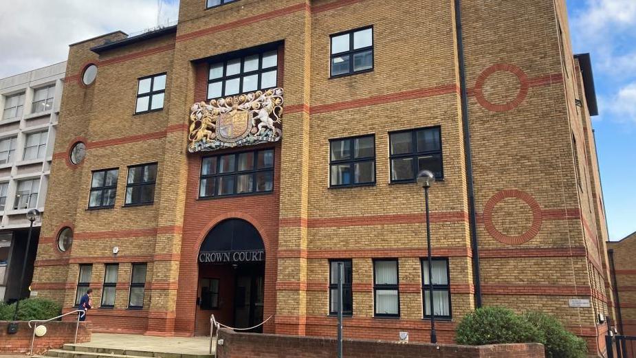 A building made of brown and red bricks. The words "Crown Court" are written over the entrance, beneath a crest.