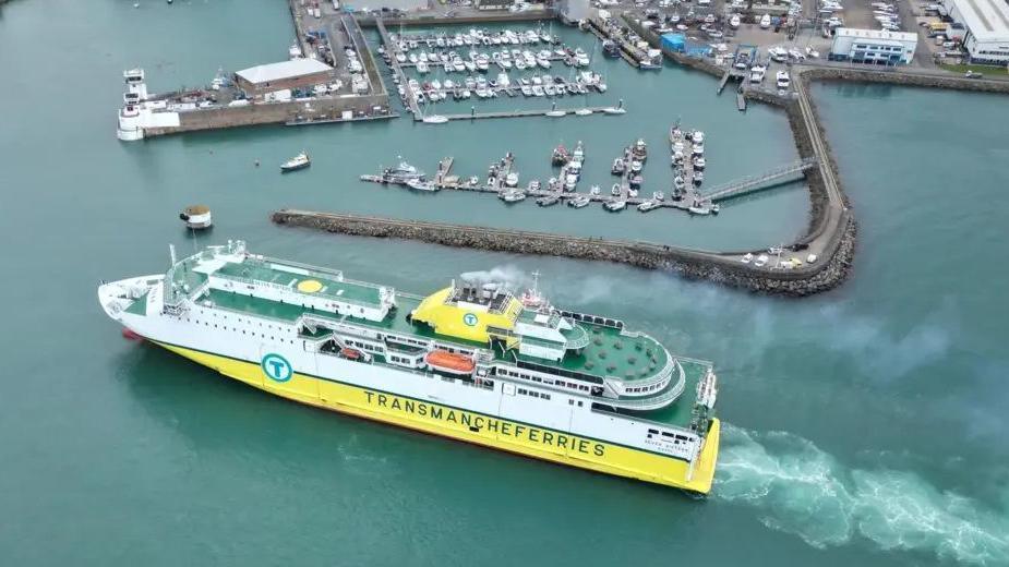 A yellow and white DFDS ferry with green decks is pulling into harbour. It has smoke coming out of its funnel and is leaving a wake. On its port side are the letters TRANSMANCHEFERRIES.