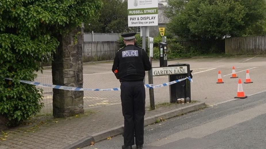A police officer is seen standing next to a car park with his back to the camera. There are bollards around the area and police tape.