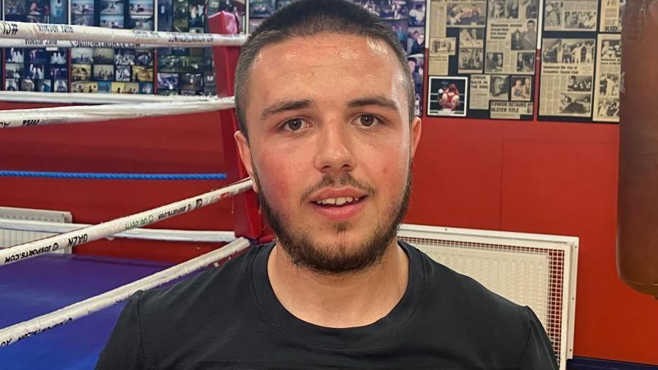 22-year-old Ryan Johnson at Monkstown Boxing Club, he is wearing a black T-shirt and standing in front of a boxing ring