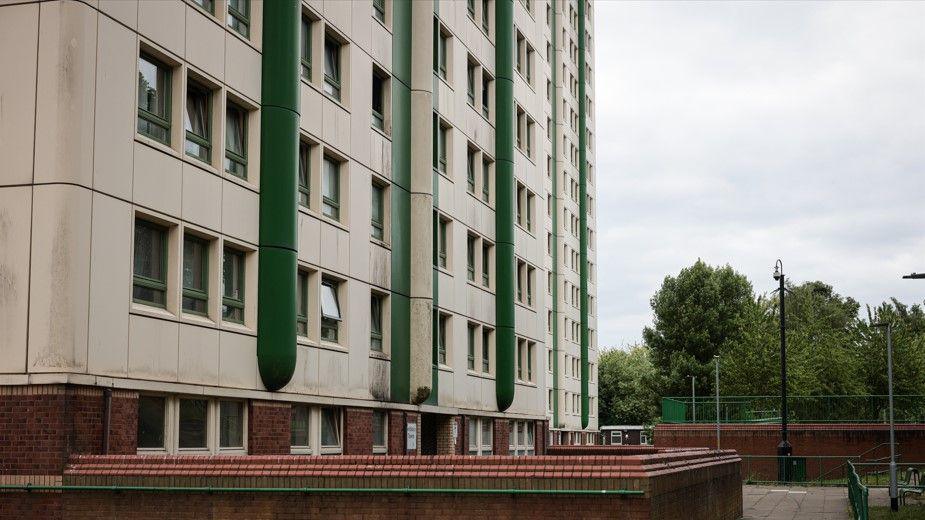 A tower block is white and green. A brick wall with a handrail can be seen outside, along with a paved area and trees.