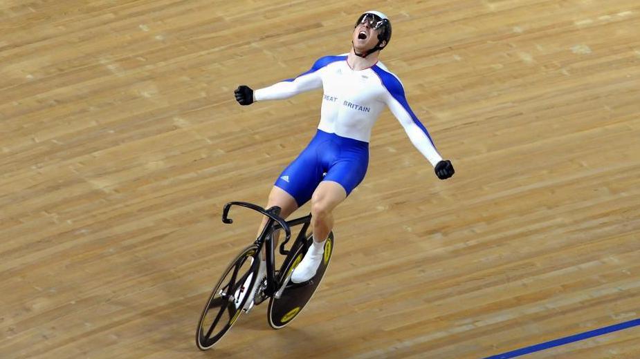 Chris Hoy in white and blue kit with his arms outstretched victoriously as he rides a bike