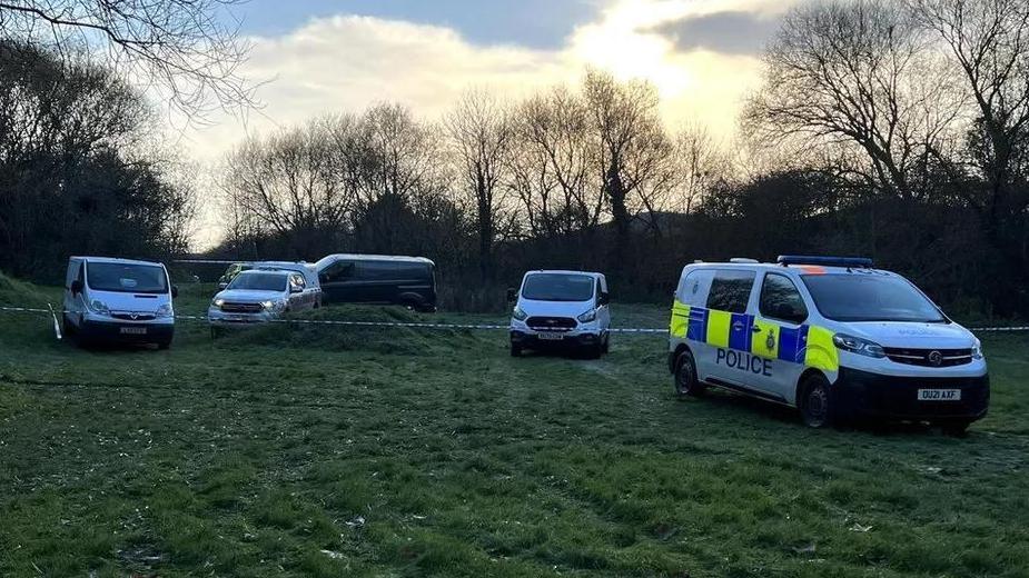 At least six police vehicles sit on grassy land on Brackley Park, there is a police cordon in place by the vehicles.