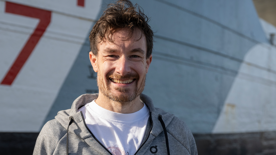 Chris Thompson, smiling, with a grey hoody and white T-shirt, standing in front of a grey D-Day landing craft on Southsea seafront