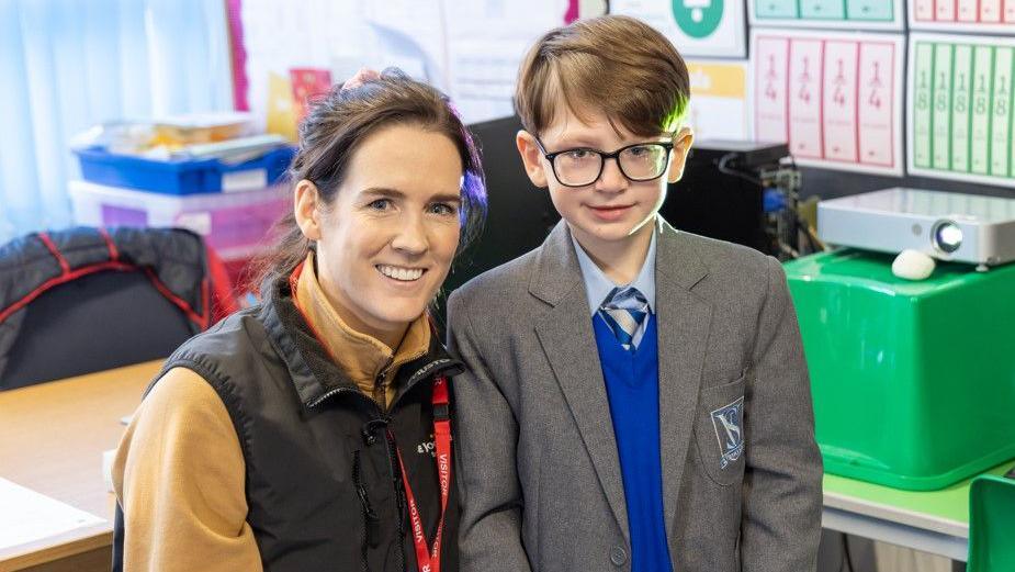 Jockey Rachael Blackmore is crouched next to Joshua in a school classroom. Rachel has brown hair tied back and is wearing a beige fleece with a black gillet. Joshua had short brown hair and is wearing dark framed glasses and a grey school uniform with blue jumper and blue and grey stripy tie. 