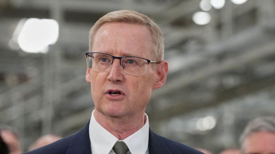 A close up shot of a man wearing glasses who is looking to the left of the frame and appears to be mid speech. He is wearing a blue suit and white shirt which are barely visible at the bottom of the photo