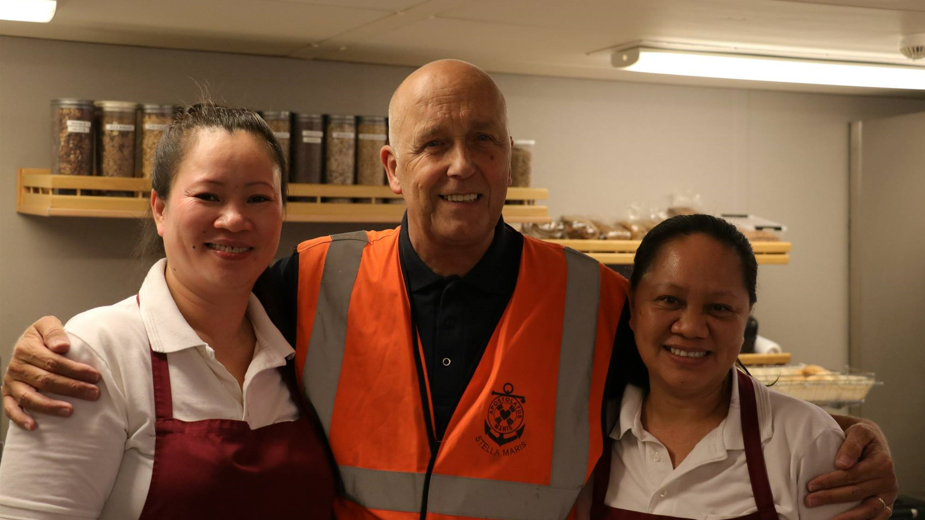 Paul Atkinson wearing a high-vis jacket standing in a kitchen with his arms around two women in aprons. 