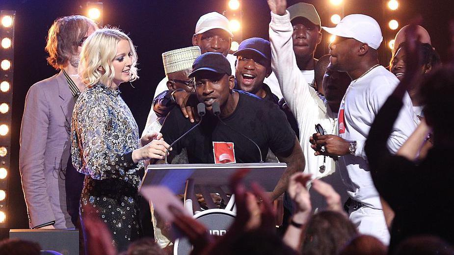 Skepta, wearing a black t-shirt and black cap, on stage accepting the Mercury Prize. He is surrounded by friends and family
