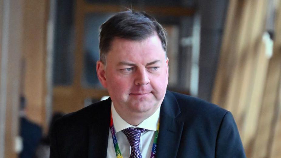 Colin Smyth, a man with flat brown hair, while wearing a suit, white shirt and purple tie and multicoloured lanyard