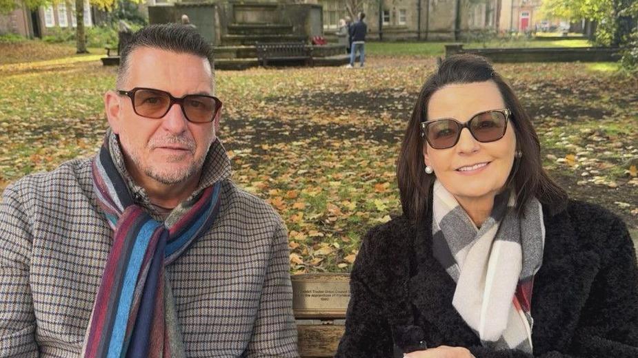 A man and a woman sit on a wooden bench in a park during autumn. The ground is covered with fallen leaves, and trees with green and yellow foliage frame the background. Both wear sunglasses and scarves. The man on the left wears a checked coat with a multicoloured scarf; the person on the right wears a black coat with a light scarf.