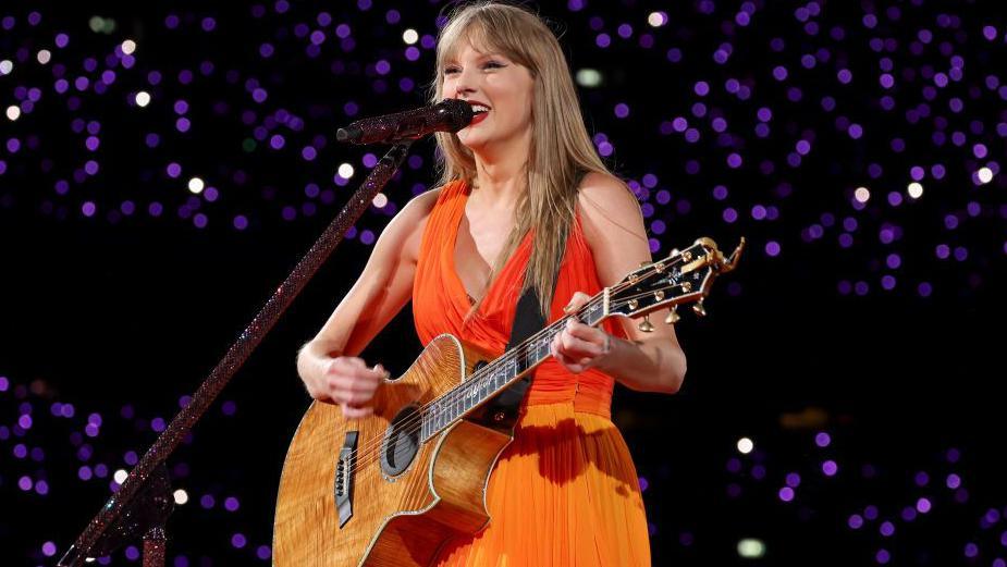 Taylor Swift wearing an orange dress and holding a guitar on stage.