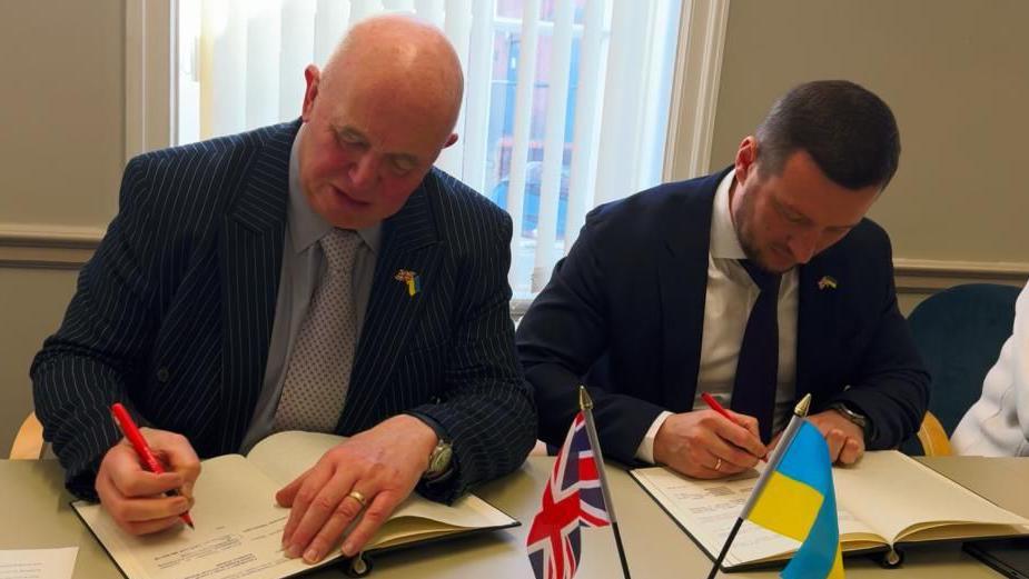 A photograph of Councillor Colin Davie and Oleksandr Prokudin bent over a desk, both signing documents of the agreement