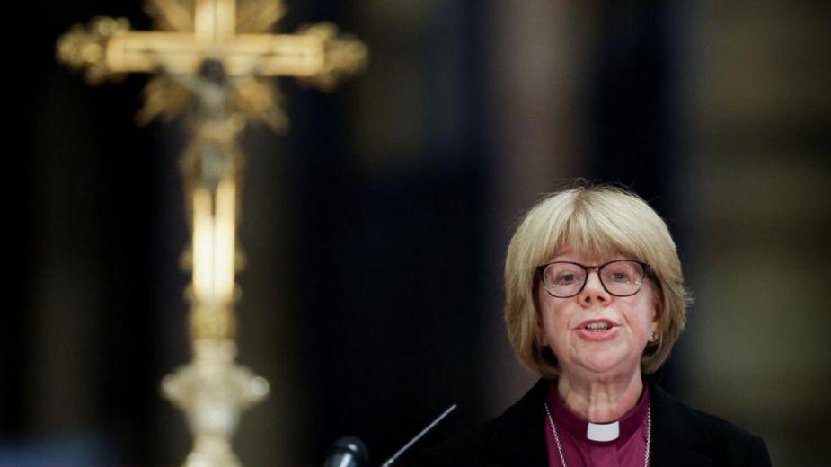 Dame Sarah Mullally, the new Archbishop of Canterbury designate, is pictured at Canterbury Cathedral speaking into a microphone with a large golden crucifix on an altar behind her. 