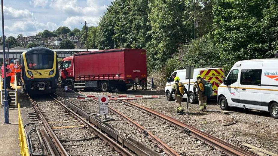 A train with a lorry parked next to it on train tracks. Network Rail vans and people in hi-vis jackets are on the scene