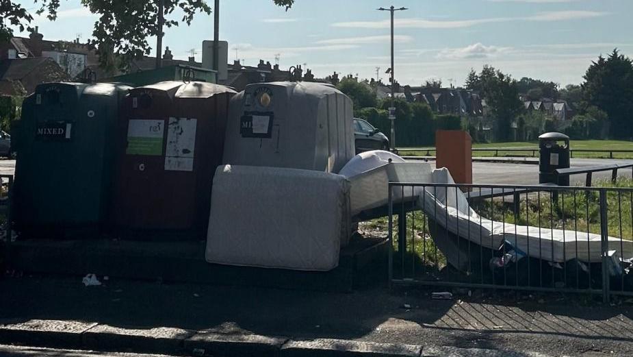 A mattress and a bed frame dumped next to a row of large recycling bins in a public car park.
