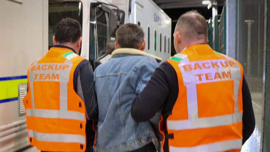 Two men in orange high-viz vests with back-up team written on the back hold a man in a denim jacket by the arms as they take him onto a police van