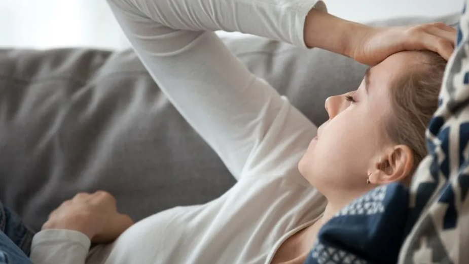 A woman lying on settee with her hand on her forehead. She is wearing a white top and lying on a blue and white cushion