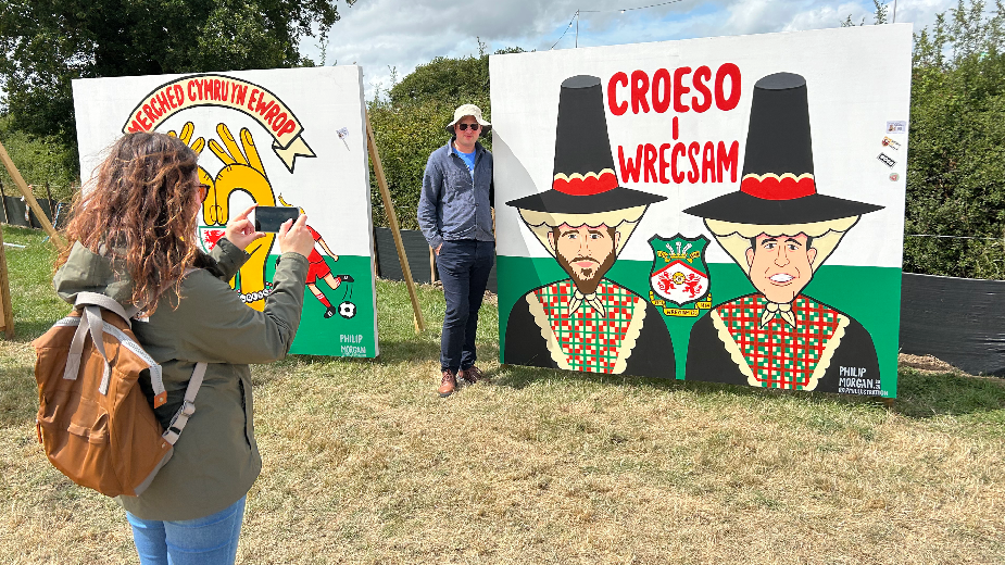 A woman taking a picture of a man next to a giant mural of the faces of Ryan Reynolds and Rob McElhenney wearing tall Welsh hats 