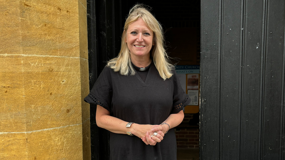 A vicar wearing a black robe, clasping her hands together and smiling at the camera in front of tall church doors