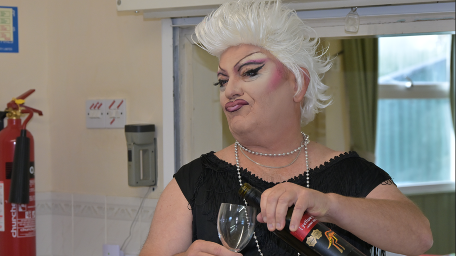 Man in drag, he has pink and white eyeshadow and strong lips along with a white wig. He wears a black top, a pearl-like necklace and is pouring a glass of red wine.