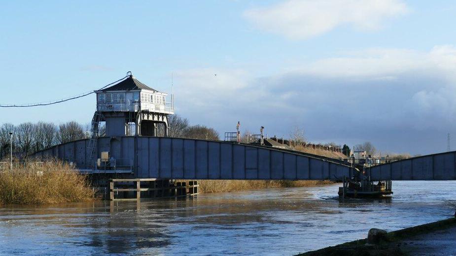 A bridge with a tower at one end spans part of the river below. It has build up sides and is made of metal.