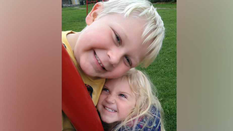 A young boy with blonde hair smiling at the camera, while a shorter, young girl with blonde hair is grinning up at him as she hugs him. There is grass behind them. 