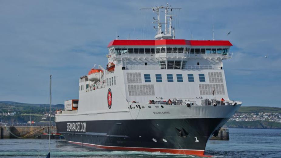 The Ben-my-Chree turning in Douglas Harbour. It is painted in the Isle of Man Steam Packet Company's colours of white, red and black and has the firm's logo featuring the three legs of Man in a red circle on the side, along with Steam-packet.com in white lettering.