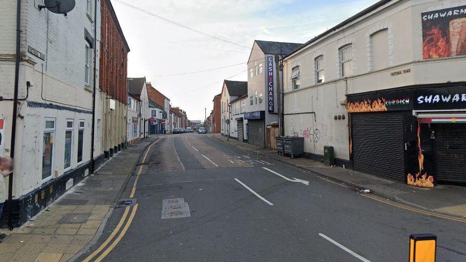 A view of Victoria Road with shops, some with shutters, and a traffic bollard in the foreground and terraced houses and cars in the distance.