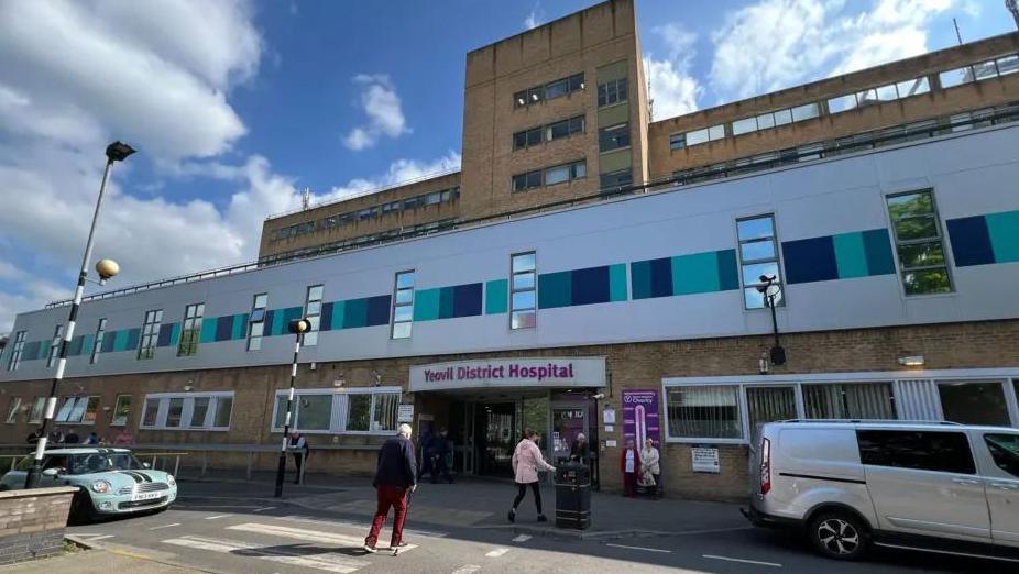 An image of a large hospital building. A sign reading Yeovil District Hospital sits over the main entrance. A number of people are crossing a zebra crossing to reach the building.