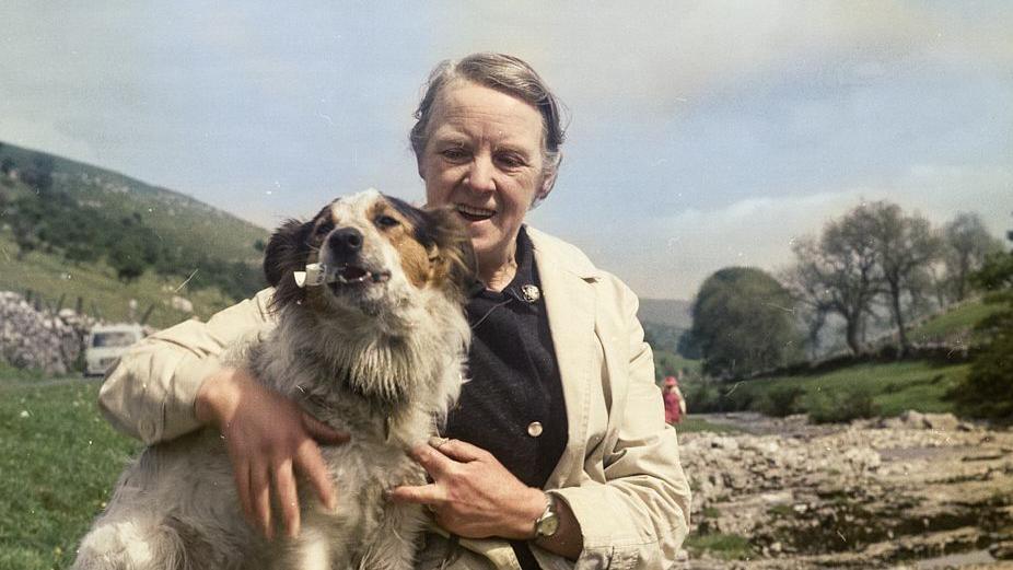 A woman with grey hair on a moor wearing a mac and a black cardigan hugs a white border collie dog.