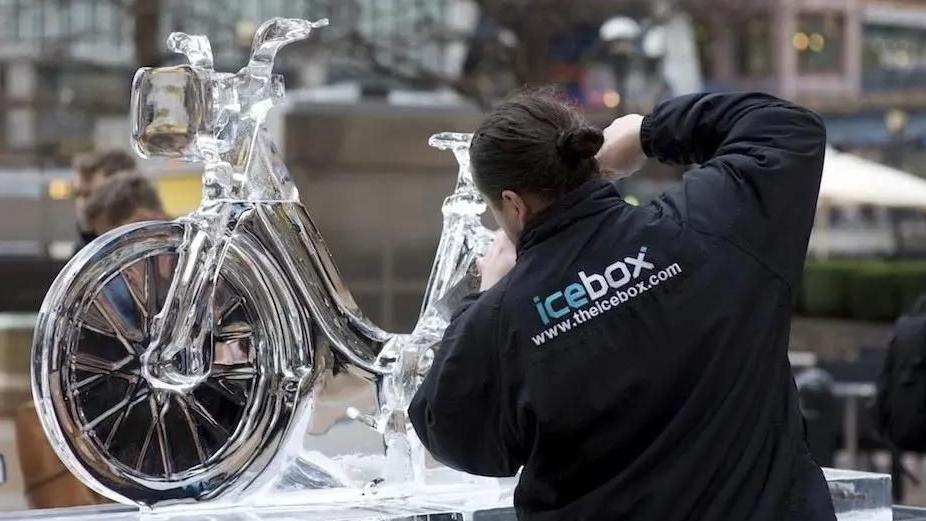 An ice sculptor working on one of last year's sculptures of a bicycle. She has her back to the camera as she works. She has dark hair and a  black jacket with the Icebox logo.