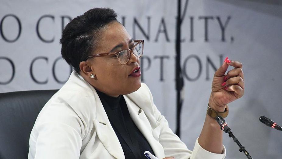 Brig Rachel Matjeng dressed in a white jacket and dark top underneath speaks into a microphone. Behind her, out of focus, is the banner for the Madlanga Commission.