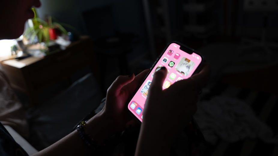 A teenager sat in a dark room using a mobile phone to access social media. The iphone has a pink background and is the focus of the photo, only the teenagers hands and arms can we seen