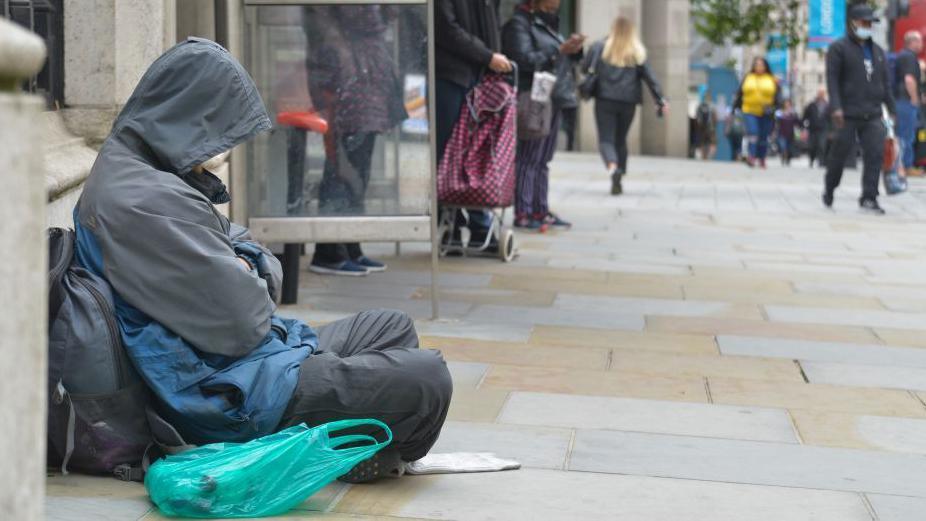 File image showing a person wearing a grey coat and trousers sleeping by a bus stop on a street near Charing Cross Station 