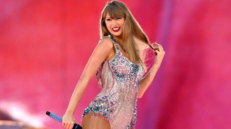 Taylor Swift holds a microphone, wearing a sparkling body suit set against a pink backdrop