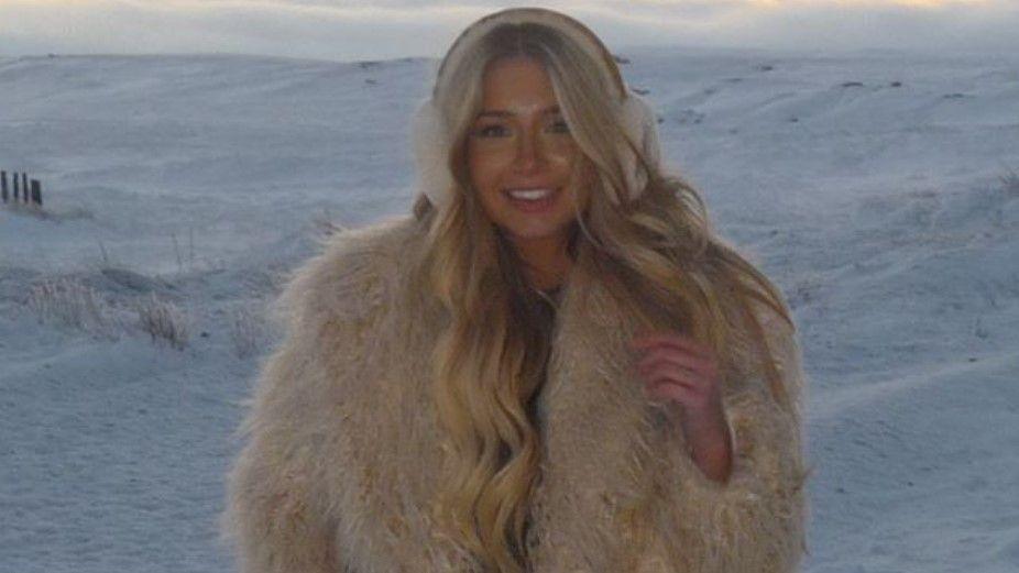 Elle Applegarth smiles at the camera as she stands in the snow. She has long blonde hair and is wearing ear warmers and a furry beige coat.