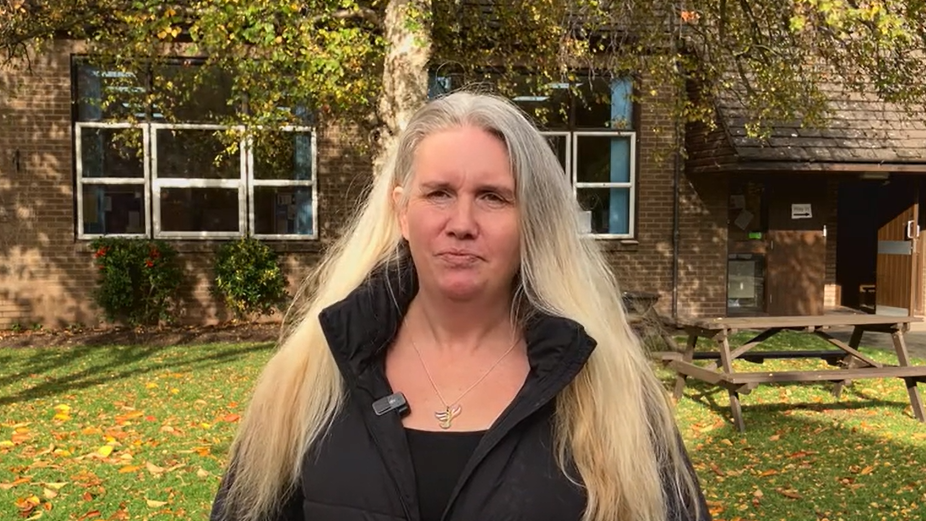 A woman with long blonde hair is pictured outside a red brick building while wearing all black clothing. 
