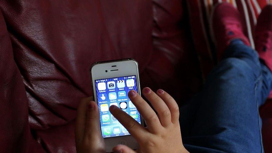 A child's hand with rough red nail varnish pointing towards an icon on a mobile phone. The child is wearing red trainers and is sitting on a brown chair.