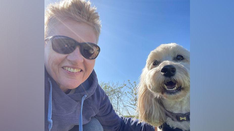 Joanne crouches alongside Murphy in a field. He has his mouth open panting a little. She wears black sunglasses and a blue hoodie, and has short blonde fur.