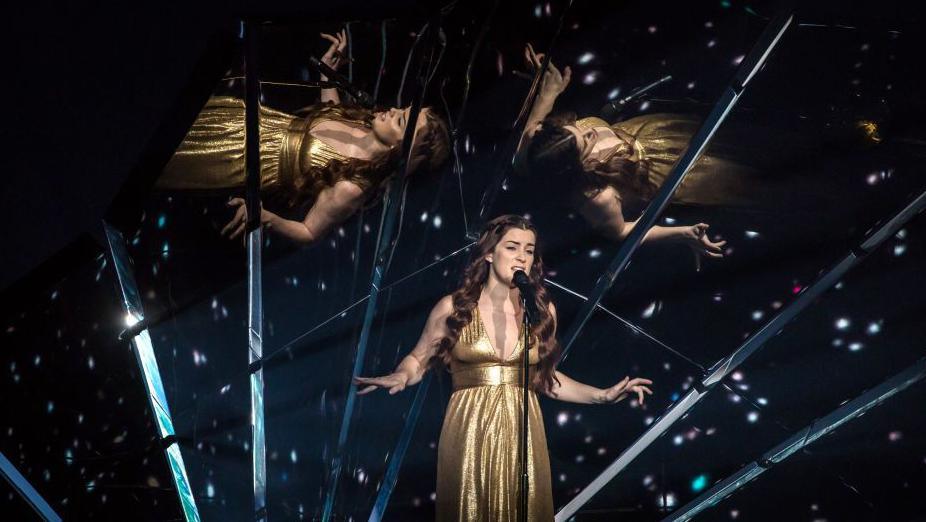 Lucy, with long brown hair, is wearing a full-length gold dress and singing into a microphone on a stand. Behind her are mirrors, with reflections of herself, and sparkly lights.