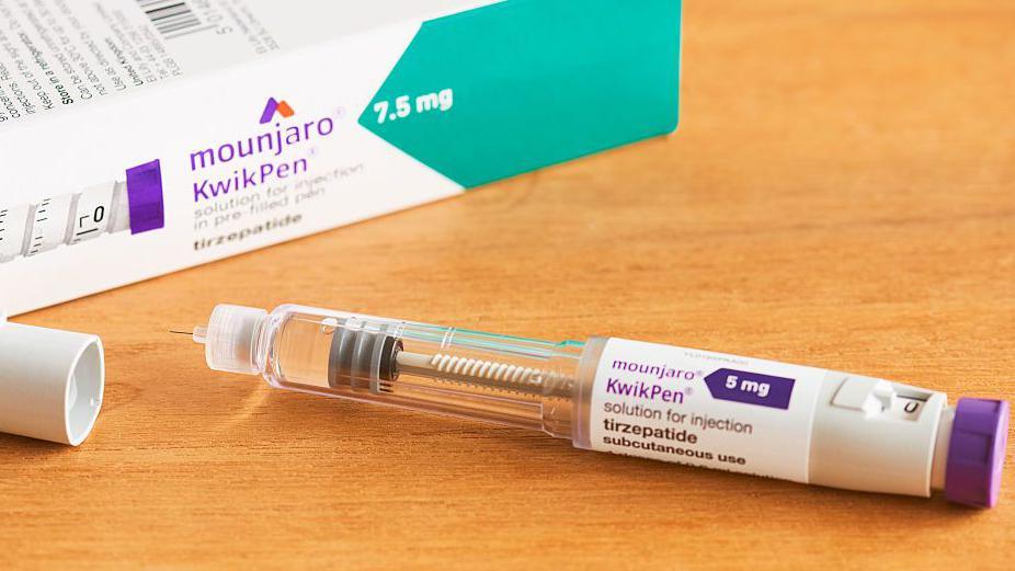 An injectable pen containing tirzepatide and branded Mounjaro KwikPen lies on a wooden table with its cap off, next to a branded box containing the injection.