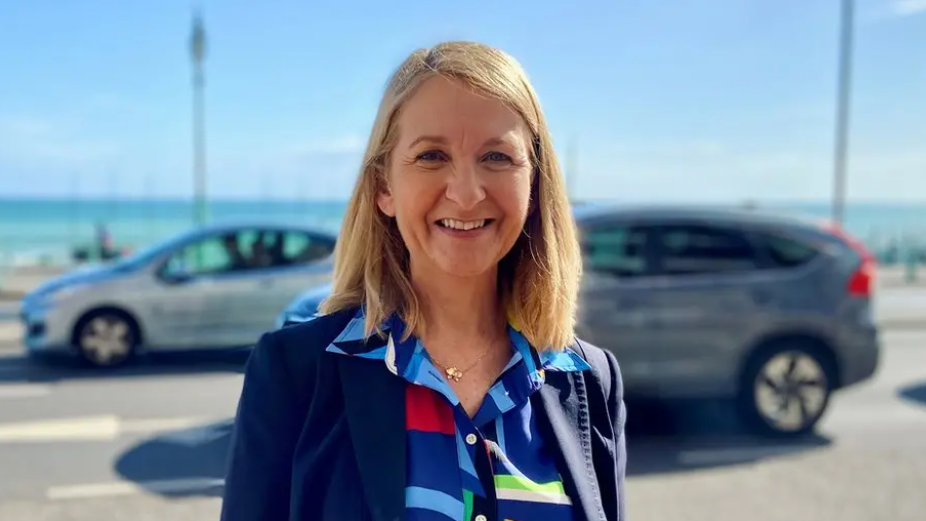 A woman smiles at the camera on a sunny day. She has blonde shoulder length hair and a bright blue shirt on. She is standing outside in front of a busy road with cars on.