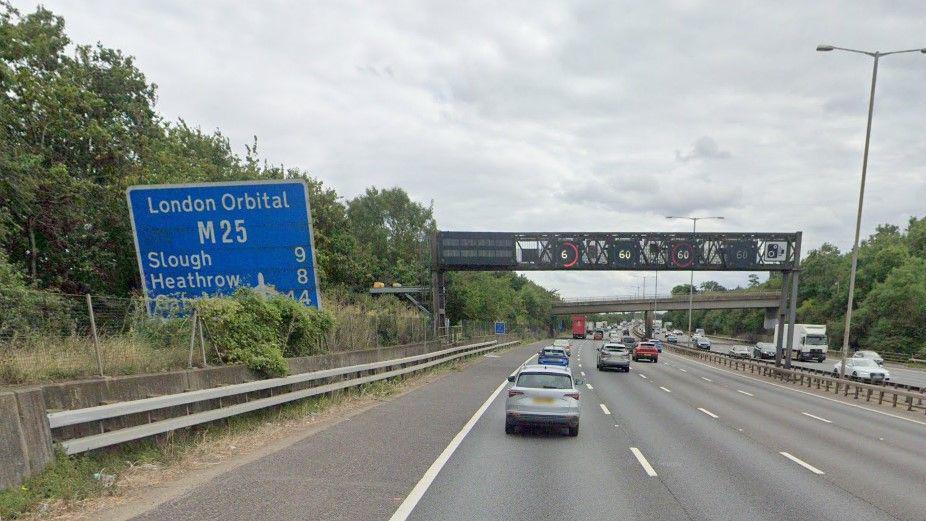 A still of the M25, with a number of cars on the road driving away from the camera. On the left is a large blue road sign which reads "London Orbital M25, Slough 9, Heathrow 8". 