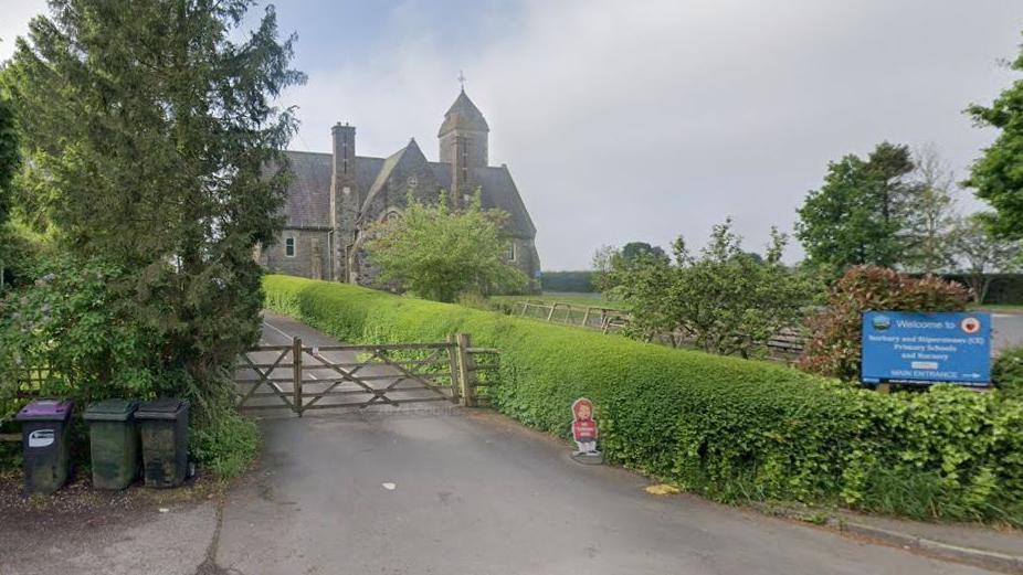 Entrance showing gate and drive to a single-storey stone-built school which is at the top of a hill in the background. It has a prominent chimney and tower with triangular roof. There is a green hedge along the side of the driveway and trees on the land between the gate and the school. There is a blue sign showing the school name.