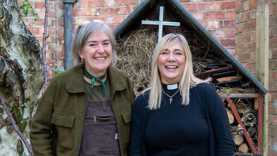 Mandy McQueenie and Rev Angie Milne standing side-by-side with a cross behind them. Mandy is wearing a green jacket with a green bow tied around her neck. Rev Angie is dressed in black with a clerical collar and a symbol of a Christian cross on a silver necklace around her neck.